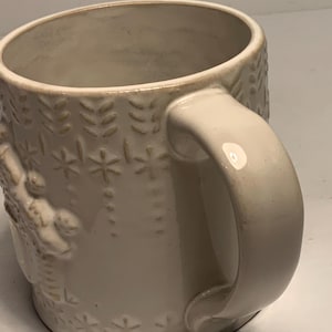 Peut inclure: Mug en c&eacute;ramique cr&egrave;me avec des motifs floraux et flocons de neige en relief. La tasse a une anse arrondie et un bord l&eacute;g&egrave;rement &eacute;vas&eacute;. Le design comprend des rang&eacute;es verticales de feuilles et des d&eacute;tails en forme d'&eacute;toiles.