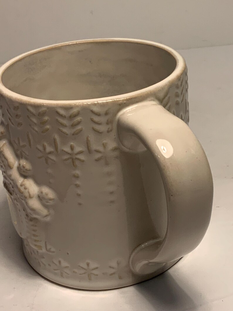 Peut inclure: Mug en c&eacute;ramique cr&egrave;me avec des motifs floraux et flocons de neige en relief. La tasse a une anse arrondie et un bord l&eacute;g&egrave;rement &eacute;vas&eacute;. Le design comprend des rang&eacute;es verticales de feuilles et des d&eacute;tails en forme d'&eacute;toiles.