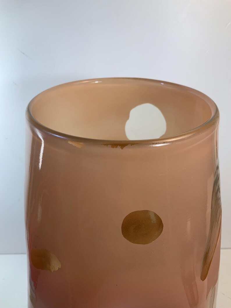 May include: A peach-colored glass vase with a cylindrical shape. The vase has a wide opening and a few brown spots on the surface. The background is white.
