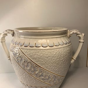 May include: A large, off-white ceramic vase with two ornate handles. The vase features a textured surface with a floral pattern and gold accents. The rim and base have decorative detailing. A classic piece for home decor.