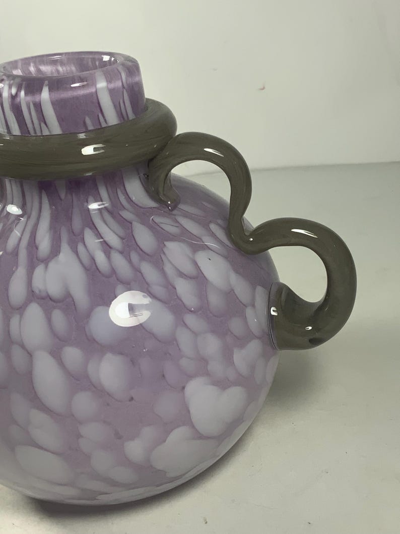 May include: A purple and white glass vase with a grey handle. The vase has a swirling pattern of white on a purple background.