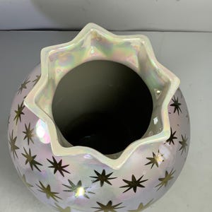 May include: A pearlescent ceramic vase with a star pattern. The vase has a round body and a scalloped rim. The stars are gold and are scattered across the surface of the vase. The rim has an iridescent sheen.