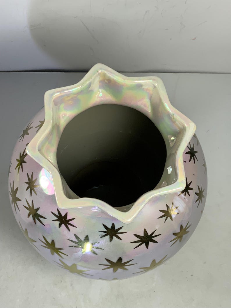 May include: A pearlescent ceramic vase with a star pattern. The vase has a round body and a scalloped rim. The stars are gold and are scattered across the surface of the vase. The rim has an iridescent sheen.