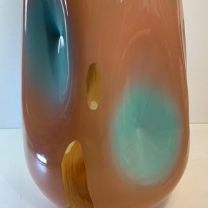 May include: A tall, rounded vase with a glossy finish. The vase features a blend of colors, including peach, teal, and gold. Teal and gold accents are layered over the peach base, creating an abstract design. The vase has a smooth, curved shape.