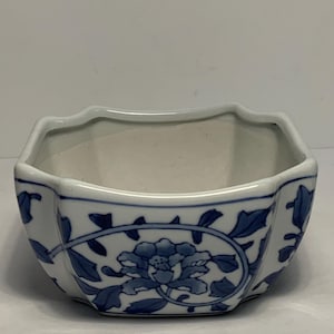 May include: A small, white ceramic bowl with a blue floral design. The bowl has a unique, octagonal shape with a scalloped rim. The blue floral pattern includes a large flower and leaves, creating a classic, decorative piece.
