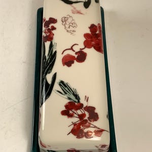 May include: A rectangular butter dish with a white lid featuring a floral design in red and black. The base is a dark teal green. The floral pattern includes stylized flowers and leaves.