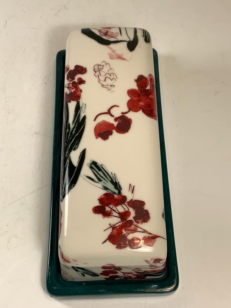 May include: A rectangular butter dish with a white lid featuring a floral design in red and black. The base is a dark teal green. The floral pattern includes stylized flowers and leaves.