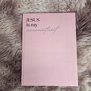 May include: A pink notebook with the text "JESUS is my accountant" in black. The notebook is rectangular and has a smooth surface. The background is a fluffy, light brown material.