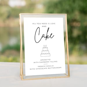 May include: A framed sign with the text "ALL YOU NEED IS LOVE AND Cake". Below the text is a line drawing of a tiered cake. The sign lists cake flavors: Amaretto with Raspberry Filling and French Vanilla with Chocolate Buttercream. The frame is gold.