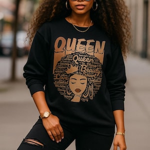 Könnte beinhalten: Schwarzes Sweatshirt mit einem goldenen "QUEEN"-Aufdruck, der das Gesicht einer Frau und Wörter wie "Love" und "Beauty" zeigt. Die Frau trägt eine Sonnenbrille, eine goldene Halskette und eine goldene Uhr. Sie trägt eine schwarze zerrissene Jeans und braune Stiefel.