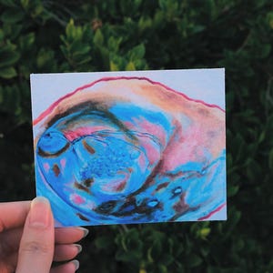 May include: A close-up of a blue, pink, and brown abstract painting on a white background. The painting has a swirling, organic shape and appears to be made with acrylic paint.