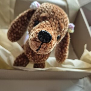 Könnte beinhalten: Ein handgefertigter brauner Häkelhund mit Schlappohren und einer schwarzen Nase. Das Plüschtier liegt in einem Karton mit Seidenpapier. Der Hund hat kleine weiße und rosa Schleifen auf den Ohren. Ein Wollknäuel ist im Vordergrund zu sehen.