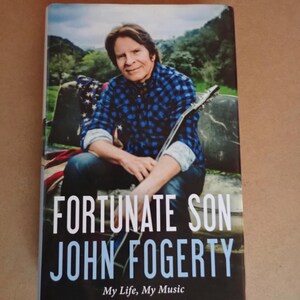 May include: A book cover featuring John Fogerty, the title "Fortunate Son," and the subtitle "My Life, My Music." Fogerty is pictured wearing a blue plaid shirt, holding a guitar, with a scenic background.