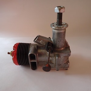 Puede incluir: Un motor de modelo vintage con culata roja y cuerpo plateado. El motor tiene un perno prominente en la parte superior y varios componentes mecánicos. El diseño sugiere un motor de avión modelo clásico o antiguo.