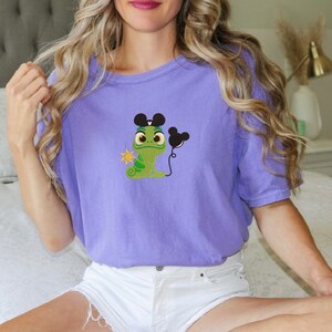 Peut inclure: T-shirt violet avec un caméléon vert portant des oreilles de Mickey Mouse et tenant un ballon Mickey Mouse. Le caméléon tient un petit soleil dans sa main.