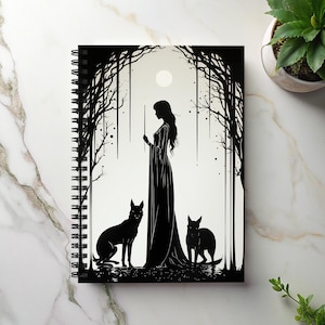 May include: Spiral-bound notebook featuring a black and white illustration of a woman in a long dress with two black dogs in a forest setting. The design includes a full moon and stylized trees. The notebook is on a white marble surface.