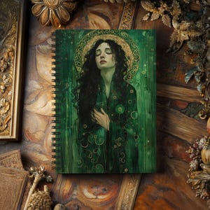 May include: Spiral-bound notebook featuring a green and gold illustration of a woman with long black hair and a golden halo. The artwork has a vintage aesthetic, with a focus on the woman's serene expression and flowing garments. The notebook is open, displaying the artwork prominently.