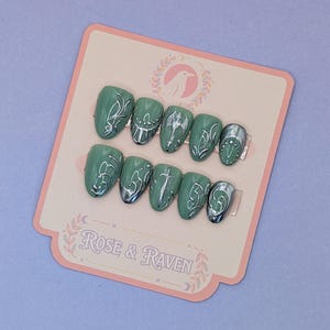 Può includere: Un set di unghie finte verde salvia con dettagli argentati. I disegni includono volute ornate, un'ascia, una croce e una luna crescente. Il testo "Rose & Raven" è su una carta rosa.