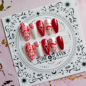 Peut inclure: Ensemble de dix faux ongles blancs et rouges avec un motif de serpent et de fleurs. Les ongles sont présentés sur une carte blanche avec le texte "ROSE & RAVEN".