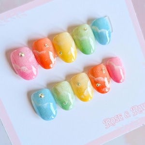 Pode incluir: Um conjunto de dez unhas postiças coloridas com as cores do arco-íris. Cada unha tem uma cor diferente, incluindo rosa, laranja, amarelo, verde e azul, com um acabamento brilhante e uma pequena joia. As unhas estão dispostas em um cartão branco com o texto "Rose & Rav" em rosa.