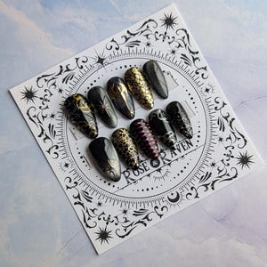 Puede incluir: Un conjunto de diez uñas postizas negras y doradas con varios diseños, incluyendo una serpiente, una estrella y una luna creciente. Las uñas están dispuestas en un círculo sobre un fondo blanco con un diseño en blanco y negro. El texto "Rose & Raven" es visible en el centro del círculo.