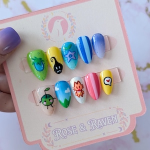 May include: A collection of ten almond-shaped press-on nails with various designs. The nails showcase cartoon characters, stripes, and abstract art. They are presented on a pink card with the text "Rose & Raven".