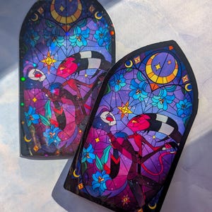 May include: Two stained glass stickers featuring a couple in a romantic pose. The stickers are in a gothic arch shape and have a purple, blue, and yellow color scheme. The couple is surrounded by flowers and stars.