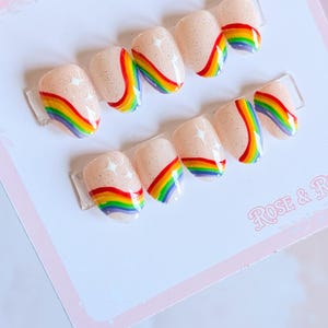 Puede incluir: Juego de uñas postizas con base nude y diseños de arcoíris. Cada uña presenta un acabado brillante y un arcoíris colorido. Las uñas están dispuestas sobre una tarjeta blanca con el texto "ROSE & R".