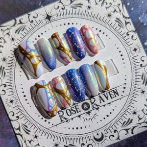 May include: Set of ten iridescent press-on nails with a celestial theme. The nails feature a variety of designs, including swirls, stars, and constellations. The nails are in shades of blue, pink, and white.