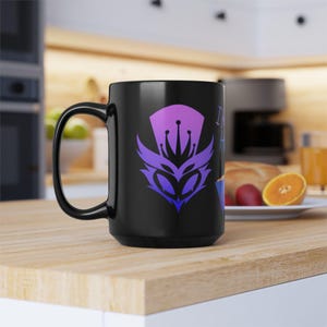 Puede incluir: Taza de cerámica negra con un diseño gráfico morado y azul de una cara estilizada y una corona. La taza tiene un asa grande y está sobre una superficie de madera. La taza es parte de una colección de artículos para beber.