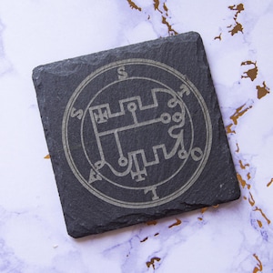 May include: A black slate coaster with a silver engraved sigil. The sigil is a circle with a complex design inside. The sigil is surrounded by a circle of text.
