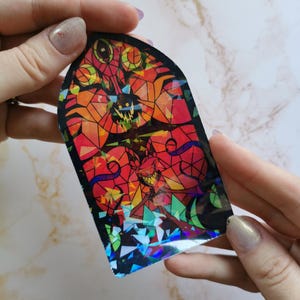 Puede incluir: Una pegatina de vitral con un dise&ntilde;o rojo, naranja y amarillo. La pegatina presenta una silueta negra de una criatura con dientes y garras afiladas. La pegatina tiene un acabado hologr&aacute;fico.