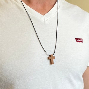 以下が含まれることがあります： 黒い紐のネックレスに木製の十字架のペンダントがついたもの。ネックレスは、赤いLevi'sのロゴが付いた白いVネックのシャツを着た人が着用しています。