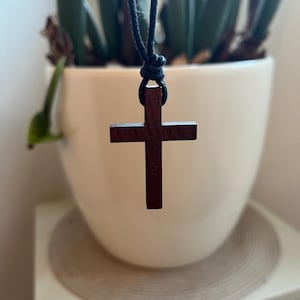 Könnte beinhalten: Ein dunkelbrauner Holz-Kreuzanhänger, der an einer schwarzen Kordel hängt. Das Kreuz hat ein schlichtes Design, und die Kordel ist oben verknotet. Der Hintergrund zeigt einen weißen Topf mit einer grünen Pflanze.
