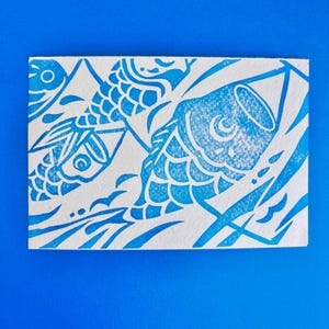 May include: A blue and white print featuring stylized koi fish. The design is a linocut style, with the fish and water elements rendered in a vibrant blue against a white background. The artwork is on a rectangular piece of paper.