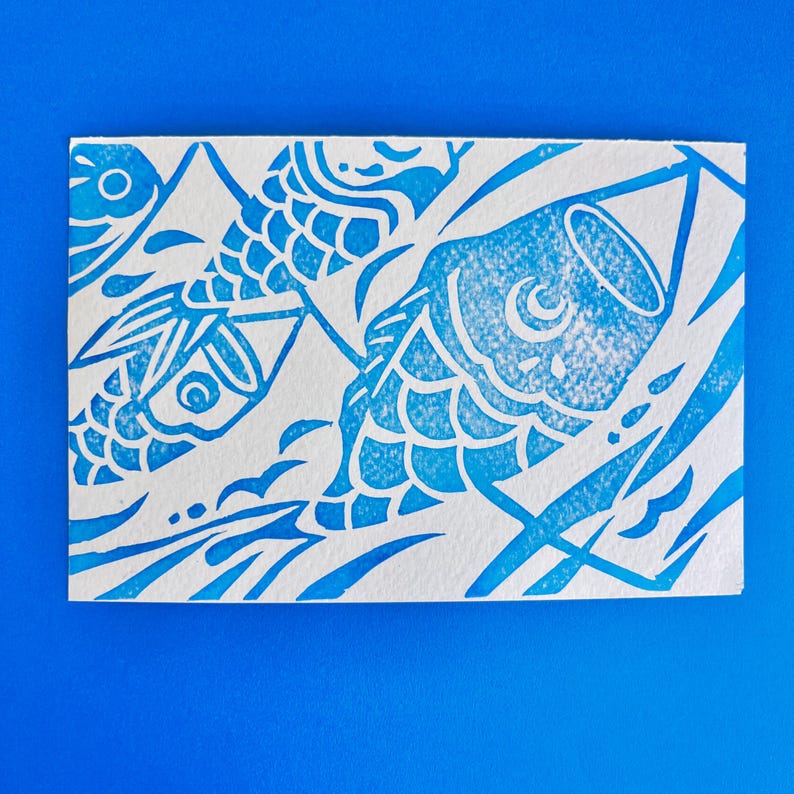 May include: A blue and white print featuring stylized koi fish. The design is a linocut style, with the fish and water elements rendered in a vibrant blue against a white background. The artwork is on a rectangular piece of paper.