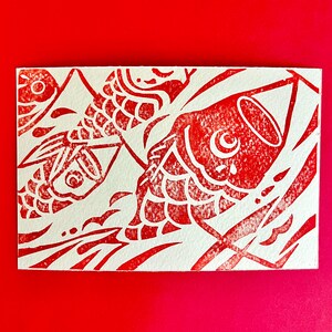 May include: A red and white print featuring stylized koi fish. The design is a linocut style, with the fish and water details in red against a white background. The artwork is on a rectangular card.