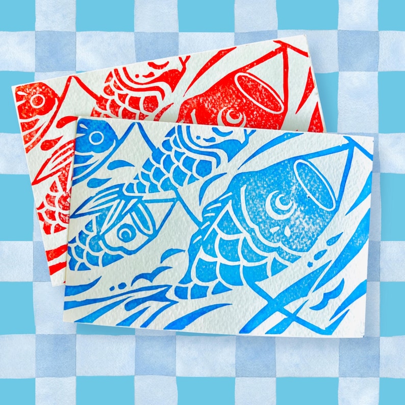 May include: Two rectangular cards with carp designs. One card features a red carp print, and the other has a blue carp print. The carp are stylized with detailed scales and fins. The background is a light blue and white checkered pattern.