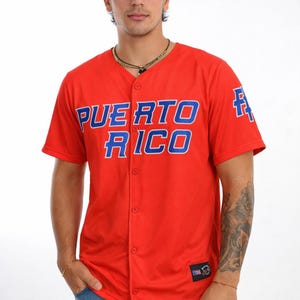 Könnte beinhalten: Ein rotes Baseball-Trikot mit den Worten "PUERTO RICO" in blauen Blockbuchstaben auf der Brust. Das Trikot hat eine Knopfleiste vorne und kurze Ärmel. Das Trikot wird mit einer blauen Jeans getragen.