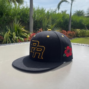 May include: Black flat-brimmed baseball cap with gold embroidered "PR" lettering. A red and green floral design is on the side, with small gold accents on the crown. The hat is displayed on a light grey surface.