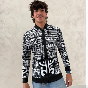 May include: Black and white zip-up jacket featuring a design with text and abstract figures. The jacket includes the Puerto Rican flag and various place names. The jacket is worn with blue jeans.