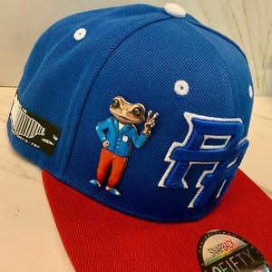 Puede incluir: Gorra snapback azul con visera roja. La gorra presenta una rana de dibujos animados con una chaqueta azul y pantalones naranjas, haciendo el signo de la paz. Detalles bordados blancos y una gran "H" blanca en el lateral.