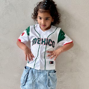 May include: A white baseball jersey with the word "MEXICO" in green and red lettering. The jersey has green and red accents and is paired with a light blue denim skirt.