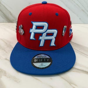 May include: A red and blue snapback hat with the letters "PR" in white and blue embroidery. The hat has a flat brim and several decorative pins. The hat is a 9Fifty style.