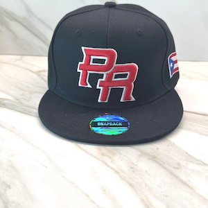 Może przedstawiać: Czarna czapka typu snapback z wyhaftowanymi czerwono-białymi literami PR z przodu. Z boku znajduje się mała naszywka z flagą Portoryko. Okrągła holograficzna naklejka na płaskim daszku zawiera napis The Original Brand Snapback of Puerto Rico.