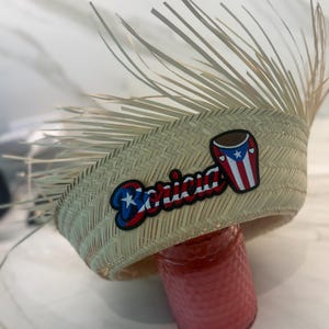 Puede incluir: Un sombrero de paja con un diseño tejido y un ala de largas y finas piezas de paja. El sombrero presenta un parche con la palabra "Boricua" en rojo, blanco y azul, junto con un diseño de la bandera puertorriqueña.