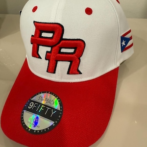 May include: White and red baseball cap featuring the letters "PR" in red embroidery. The cap has a red brim and a small Puerto Rican flag patch on the side. The cap has a snapback closure.
