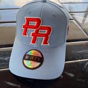 May include: Pale blue snapback baseball cap with a red and white embroidered "PR" logo. The cap has a curved peak and a "9FIFTY" sticker. The hat is made by Taino Wears.