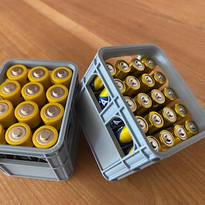 May include: Two light gray plastic crates filled with cylindrical batteries. The left crate holds twelve yellow batteries arranged in a grid. The right crate is tilted, revealing rows of yellow and blue batteries stacked inside the open-frame structure.