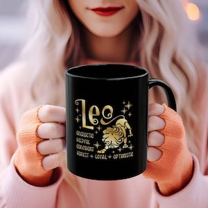 May include: Black ceramic mug featuring a gold Leo zodiac design. The design includes the word "Leo", a lion illustration, and traits such as "energetic" and "optimistic". The mug is held by a person wearing an orange sweater.